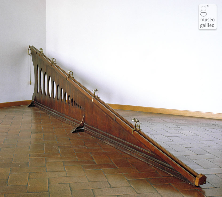 A long wooden inclined plane with holes and small bells is set up in a corner on a tiled floor at Museo Galileo.