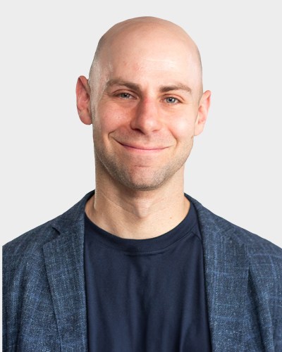 A person with a shaved head, wearing a dark blue shirt and blazer, smiles against a plain background.