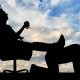 Silhouette of a selfish boss wearing a crown, relaxing on an office chair with feet propped up on another person's back against a cloudy sky backdrop.