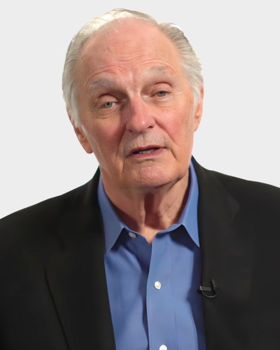 An older man in a black jacket and blue shirt is speaking, with a visible microphone attached to his shirt.