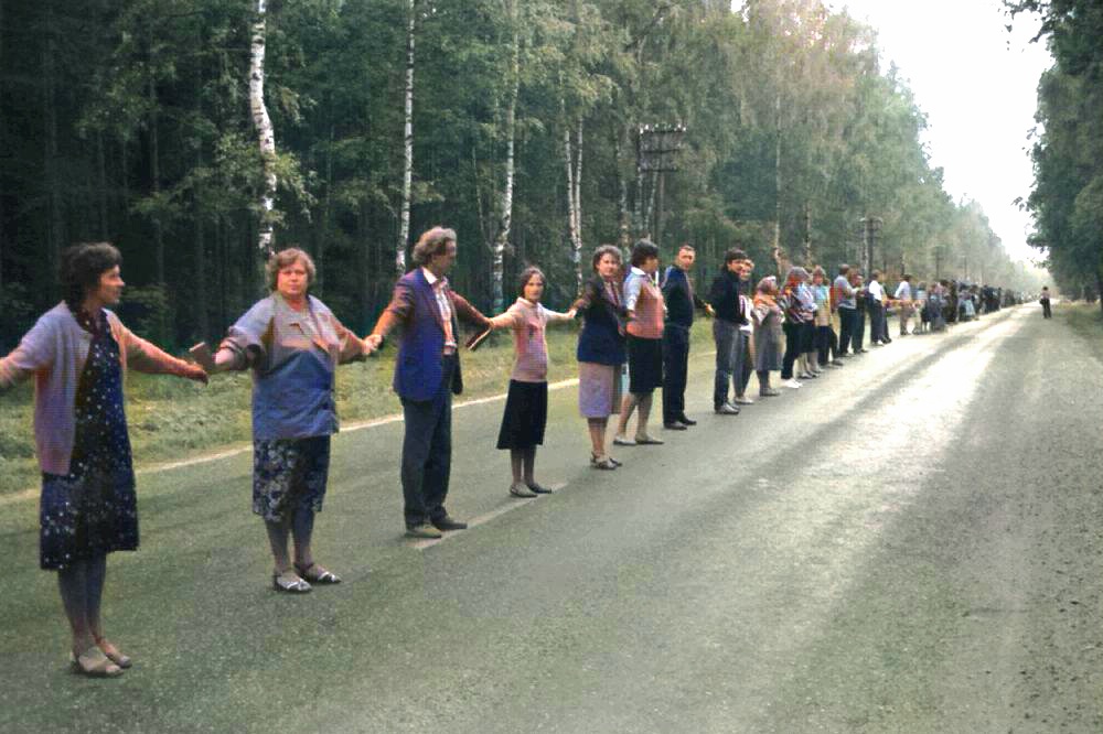 A long line of people holding hands stands on a tree-lined road, forming a human chain in a rural area.