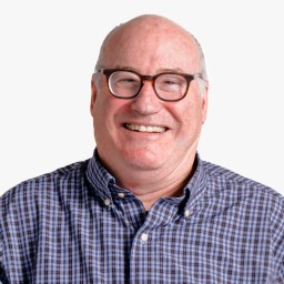 A man with glasses and a checkered shirt smiling against a plain background.