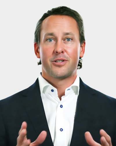 A man in a suit gestures while speaking against a plain background.