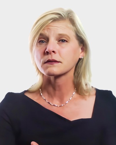 A person with blonde hair is wearing a black top and a pearl necklace, looking slightly upward against a plain gray background.