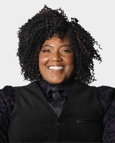 A person with curly hair smiles, wearing a black vest, dark patterned shirt, and tie against a plain background.