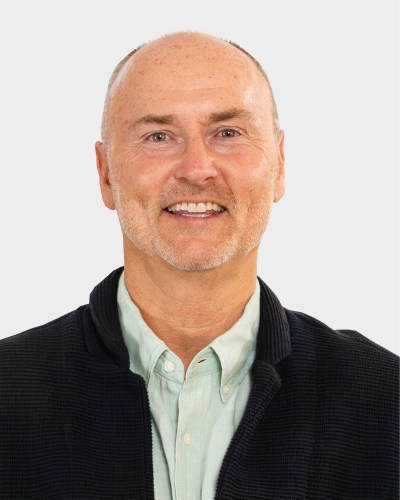 Bald man with light facial hair smiling, wearing a light green shirt and dark jacket against a plain background.