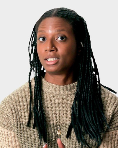 A person with long braided hair and a neutral expression is wearing a knitted sweater and a pendant necklace.