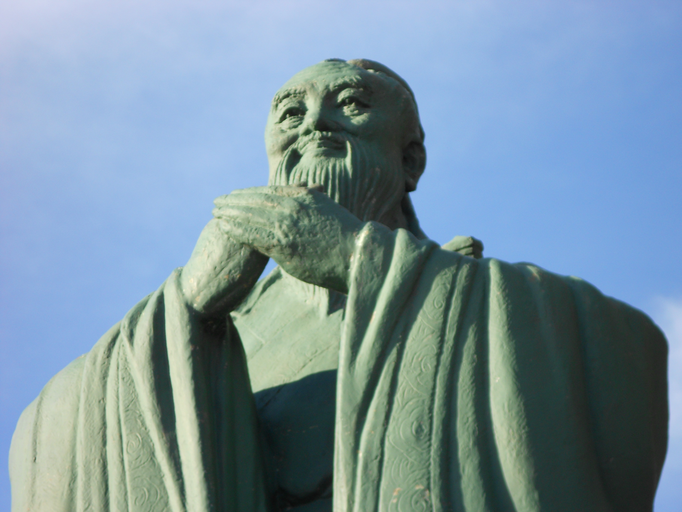 A green statue of an elderly man with a long beard and flowing robe, with hands clasped, set against a clear blue sky.