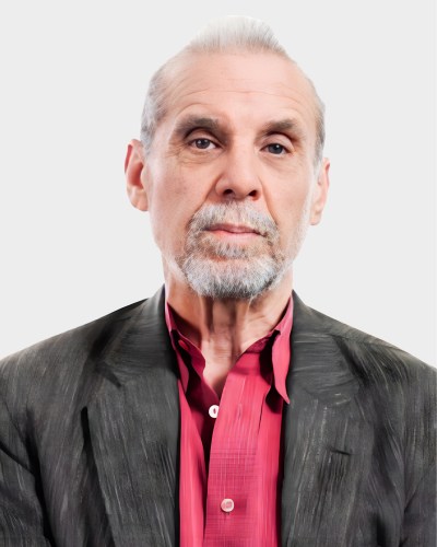 Elderly man with gray hair and beard wears a dark suit and red shirt, posing against a light gray background.