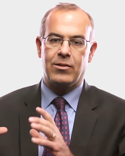 A person in a suit and tie gestures with their hands, set against a plain background.