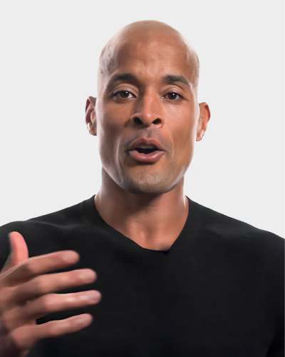 A person with a shaved head is speaking, wearing a black shirt, and gesturing with their right hand against a plain white background.