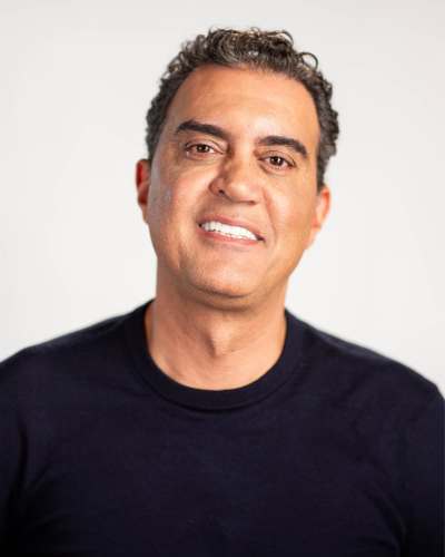 Man with short curly hair smiling, wearing a black shirt against a plain background.