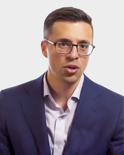 A person wearing glasses and a suit speaks or poses against a plain background.