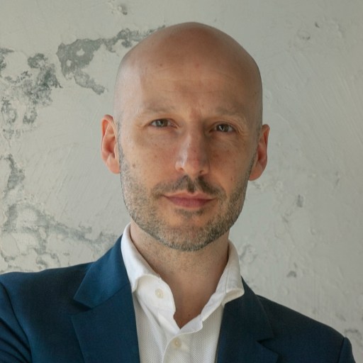 A bald man with a beard wearing a suit and white shirt stands in front of a textured wall.