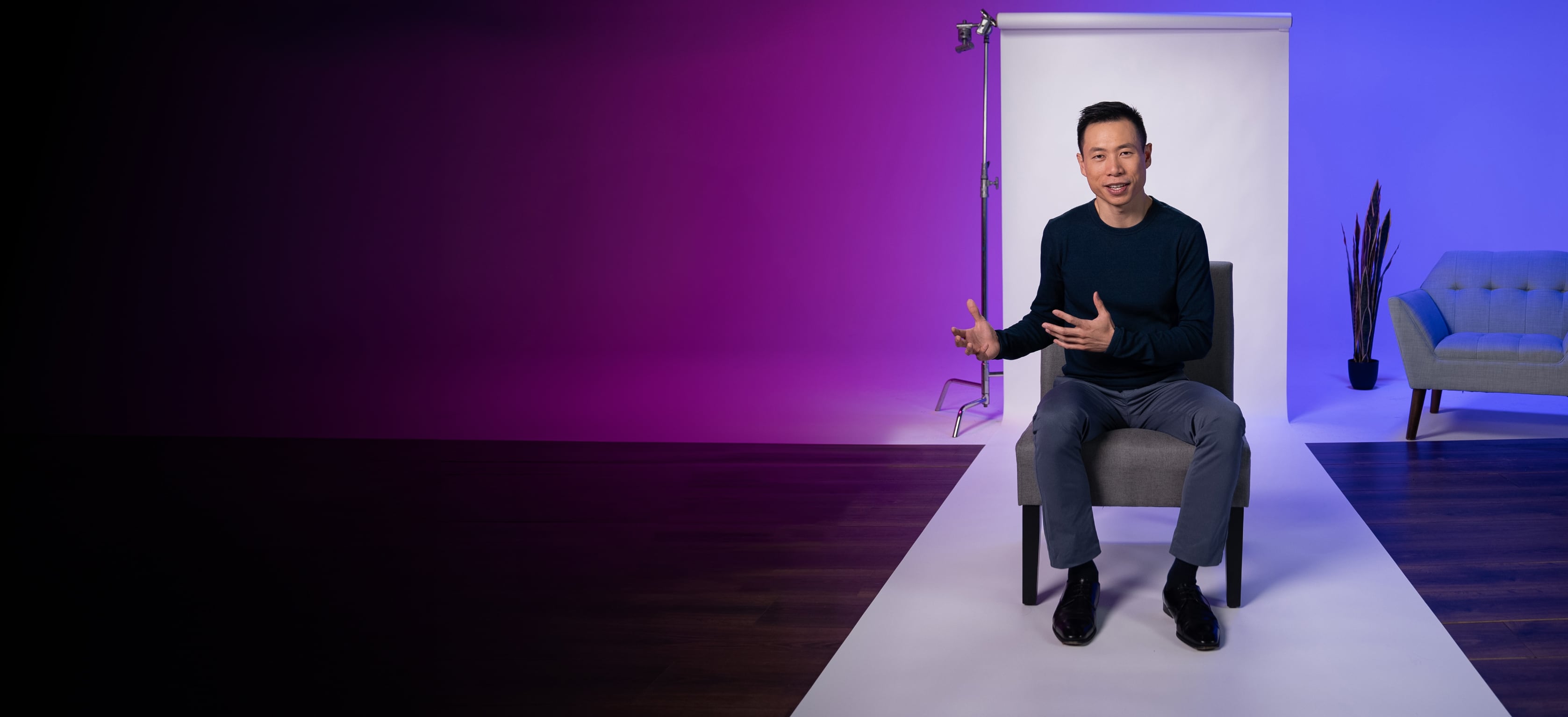 A man sits on a chair in a studio with purple and blue lighting, speaking and gesturing with his hands. A sofa and a plant are in the background.