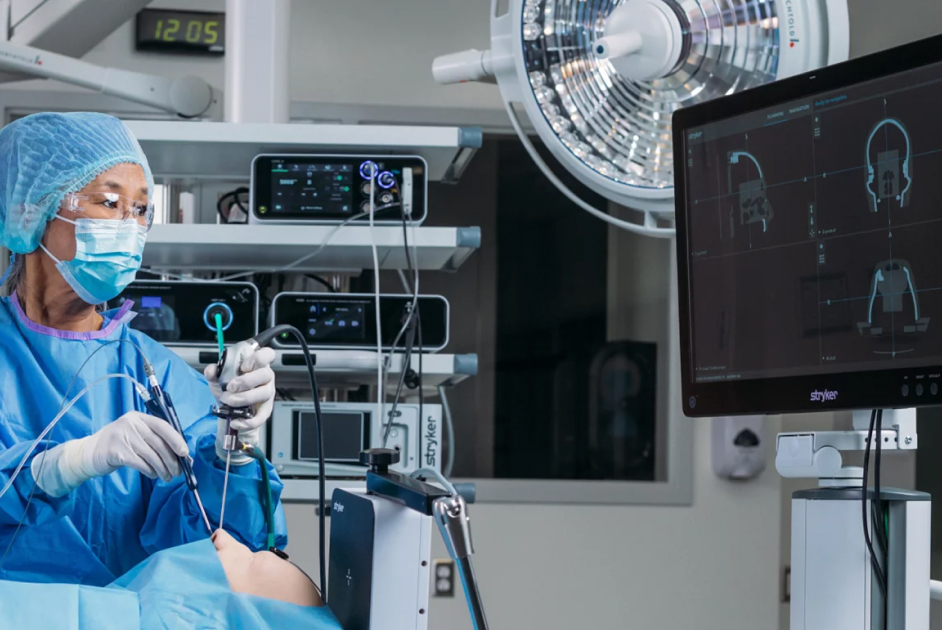 A surgeon in a blue gown and mask operates using advanced medical equipment while monitoring images on a screen in an operating room.