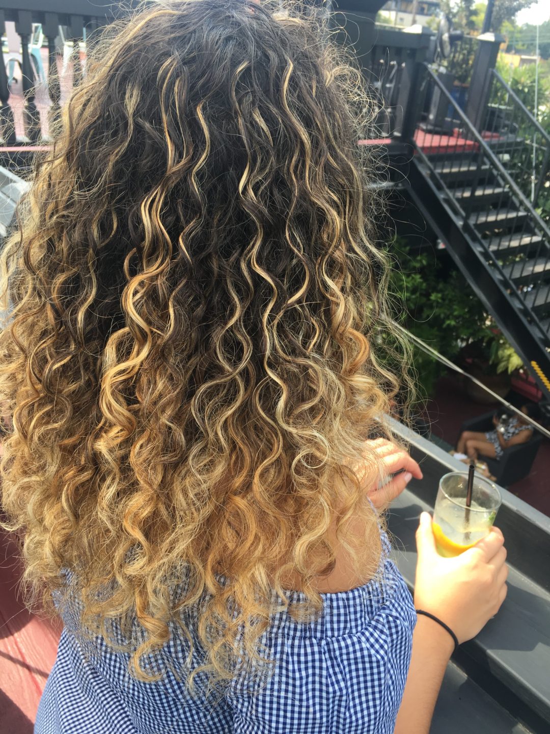 Woman with long curly hair holding a drink, standing outdoors near stairs and greenery.