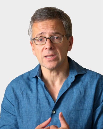 A person with short gray hair and glasses, wearing a blue button-up shirt, is speaking while gesturing with their hands against a plain background.