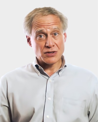 A person with short gray hair, wearing a light blue button-up shirt, looking directly at the camera with a neutral expression against a plain white background.