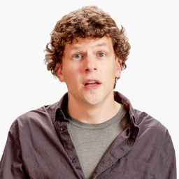 A person with curly brown hair and a neutral expression, wearing a black shirt over a gray t-shirt, stands against a plain white background.