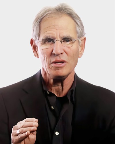 A man with glasses and gray hair is speaking, wearing a black jacket and shirt against a plain background.