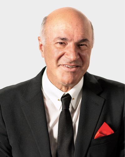 A bald man in a black suit, white shirt, and black tie with a red pocket square, posing against a plain white background.