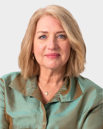 A woman with shoulder-length blond hair wears a light green satin blouse. She is looking directly at the camera with a neutral expression.