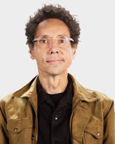 A person with curly hair, glasses, and a light brown jacket, looking straight ahead against a plain background.