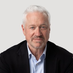 A man with white hair and a beard is wearing a dark blazer and light blue shirt against a plain background.