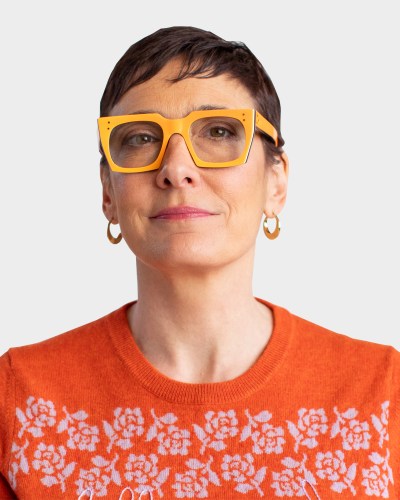 Person wearing yellow glasses and an orange sweater with floral patterns, against a plain background.