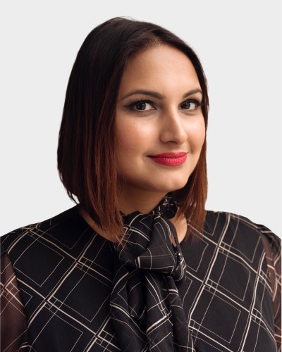 A person with shoulder-length hair, wearing a geometric-patterned black blouse, smiles at the camera against a plain white background.