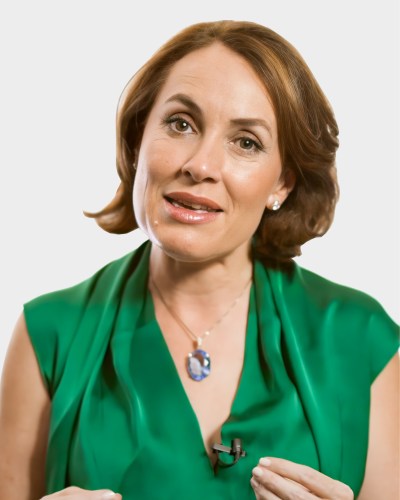 A person with short brown hair wearing a green outfit and a blue pendant necklace speaks while gesturing with one hand.