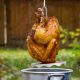 A golden-brown turkey being carefully lowered into a metal pot outdoors, ready for a deep fry amidst the grassy area and wooden fence.