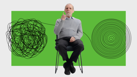 An older man sits on a chair with his finger raised. Behind him are a tangled line and a spiral, both against a green backdrop.