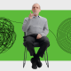 An older man sits on a chair with his finger raised. Behind him are a tangled line and a spiral, both against a green backdrop.