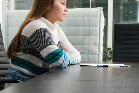 A person with long hair sits at a conference table with a document and pen in front of them.