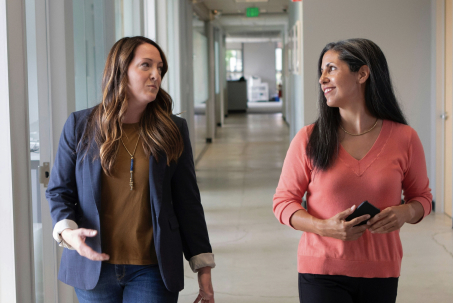 Two women walking and talking in a hallway, one holding a phone.