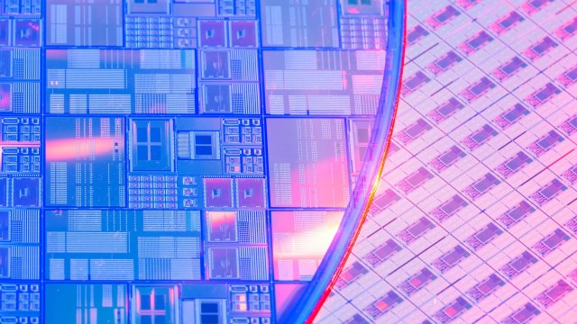 Close-up of a semiconductor wafer with intricate microchip patterns and a reflective surface, illuminated by pink and blue lights.