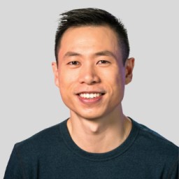 A person with short dark hair and a blue shirt smiles at the camera against a plain background.