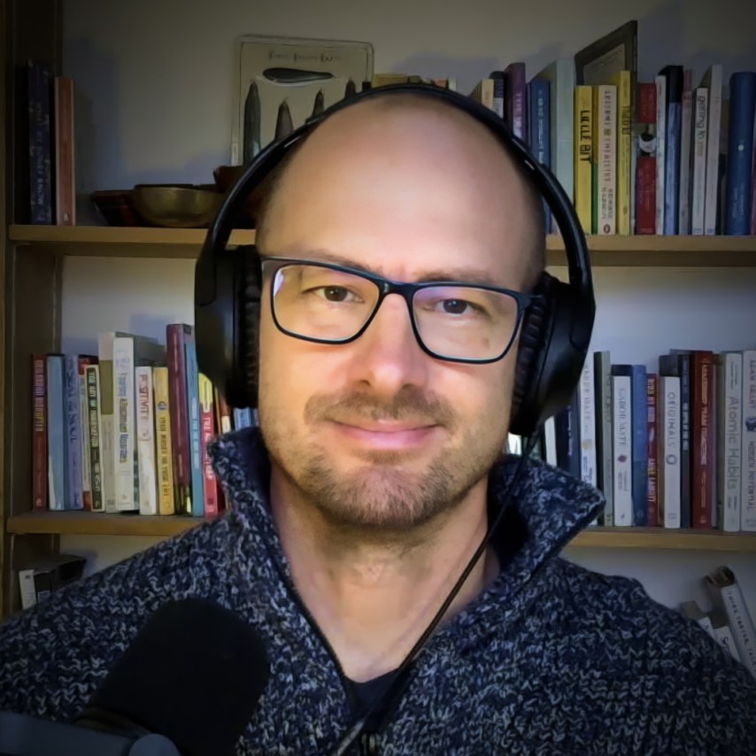 A person wearing headphones and glasses sits in front of a bookshelf, speaking into a microphone.