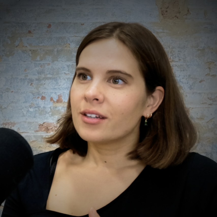 Woman with brown hair speaking into a microphone against a textured wall background.