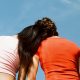 Two people viewed from behind against a blue sky, one wearing a pink top and the other an orange top, share a moment of quiet solace as the person in pink rests their head on the other's shoulder. It’s a serene scene that hints at healing and support after trauma.