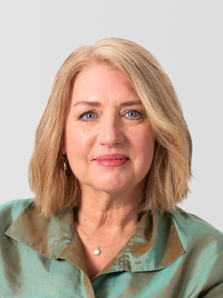 Liz Wiseman wearing a green and brown blouse and a small pendant necklace, looking directly at the camera against a plain background.