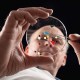 Person examining a petri dish with tweezers, holding small colored objects, against a dark background.