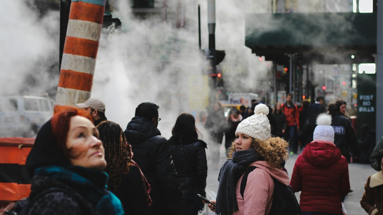 People walking on a city street with steam rising from vents create a scene reminiscent of an omics exposome research study. A woman in a white beanie looks back as buildings and traffic form the vibrant backdrop.