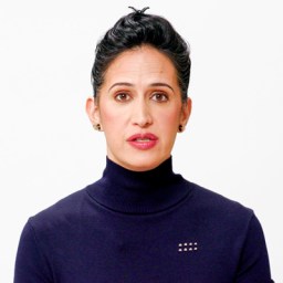 Person with dark hair in a bun, wearing a navy turtleneck, against a plain white background, looking forward.