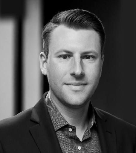 A man in a suit jacket and dotted shirt looks directly at the camera in this black and white portrait.