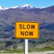 A yellow road sign reading "SLOW NOW" stands before a breathtaking mountainous landscape under a clear blue sky, reminding travelers of the art of slow productivity.