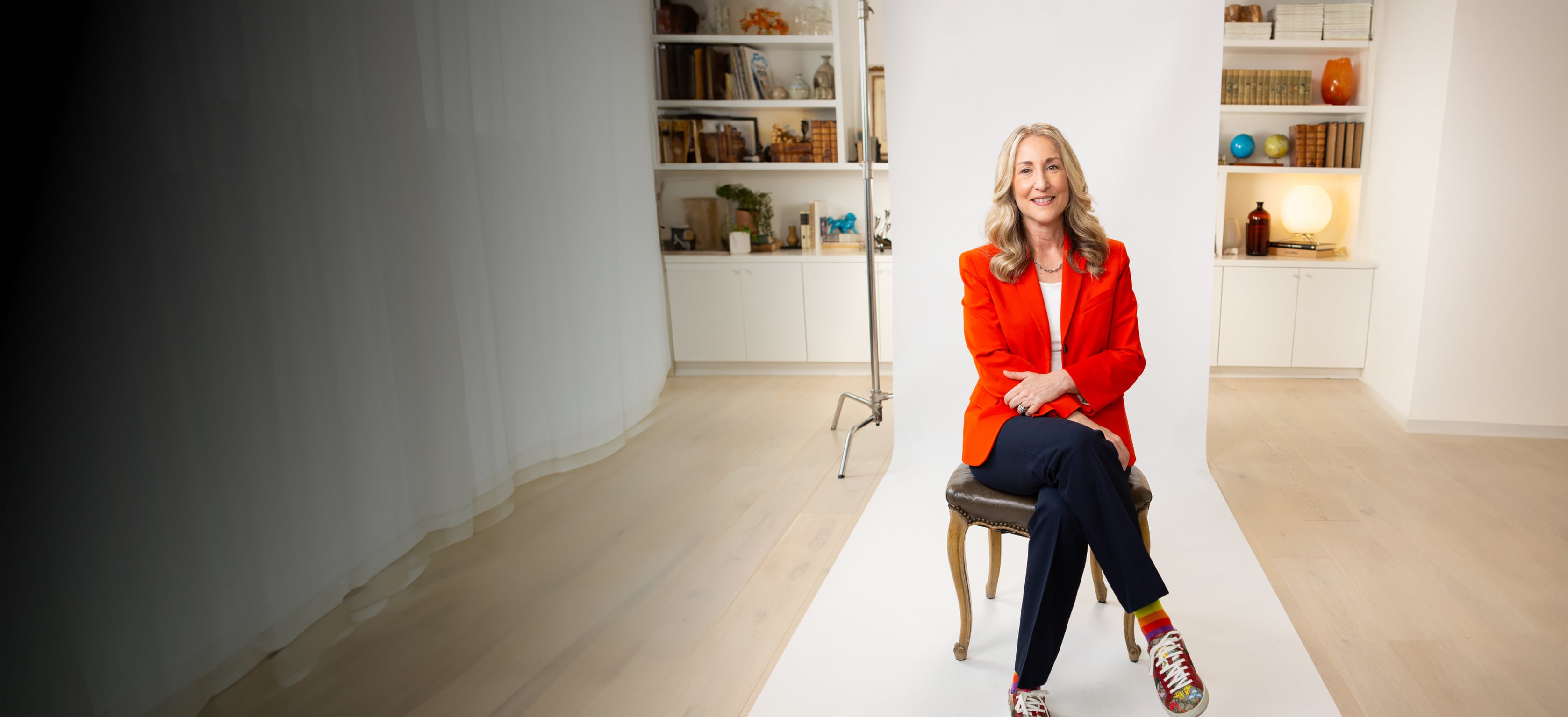 A person in a red blazer and colorful shoes sits on a chair against a white backdrop in a brightly lit room with shelves.