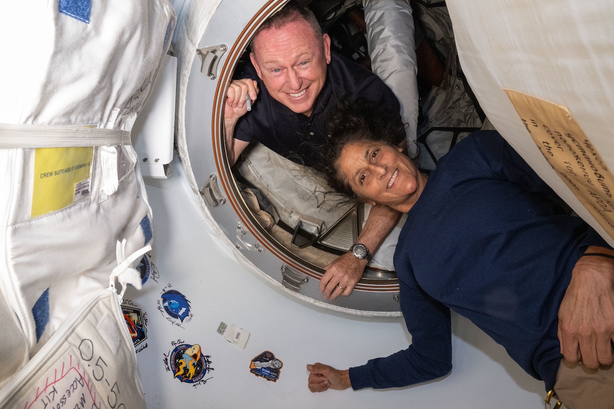 Two astronauts inside a spacecraft, one peeking through a hatch and the other floating, surrounded by mission patches.